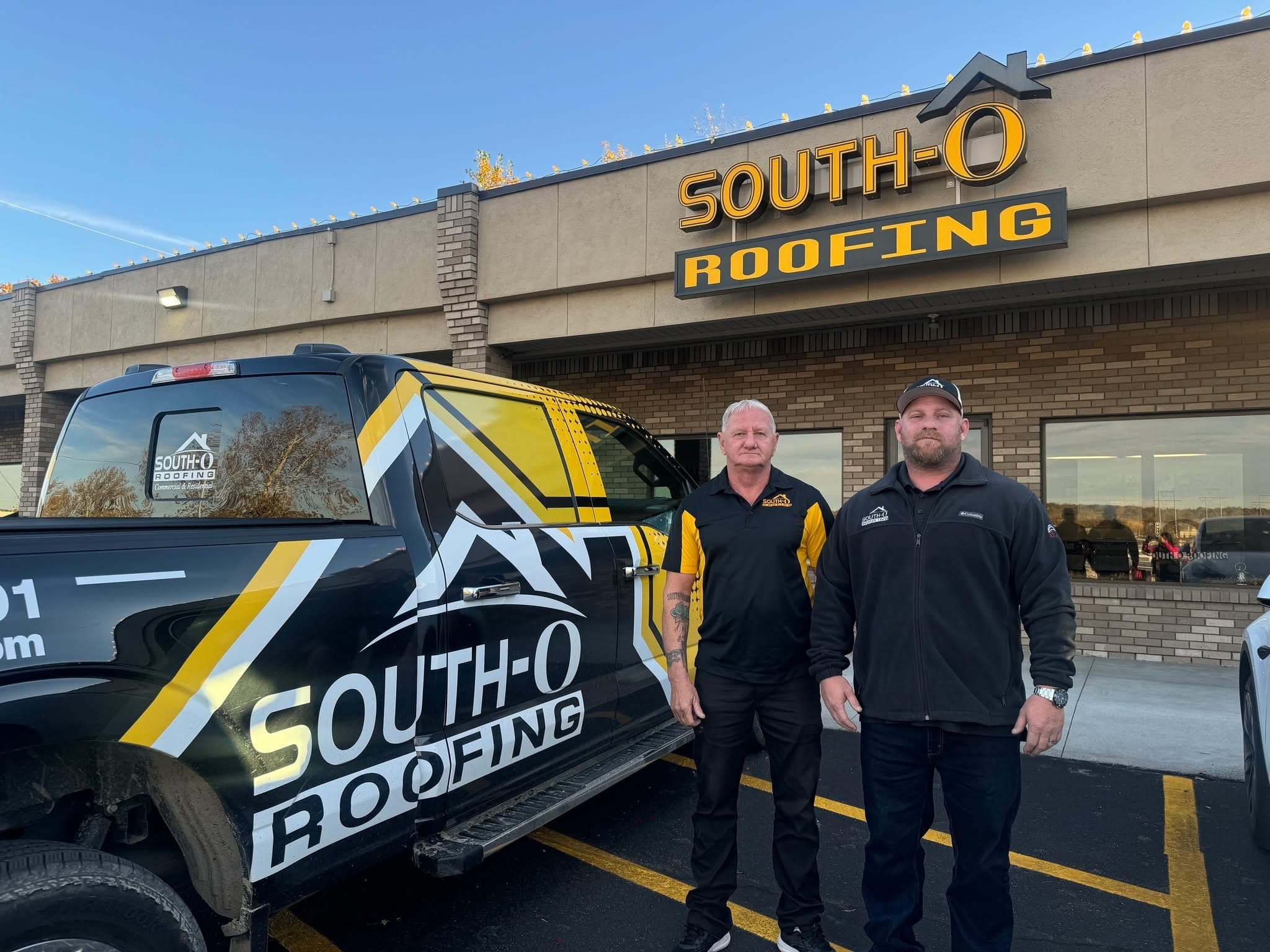 Two South O Roofing team members outside the company office, with lit South-O Roofing signage overhead and a wrapped company pickup parked out front